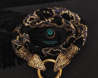 Puede incluir: Un collar de cuentas con un patrón de serpiente en negro y morado iridiscente, acabado con dos cabezas de serpiente de metal en tono dorado y un cierre circular dorado. El centro de la espiral muestra el texto STASIA BEADS beaded jewelry.