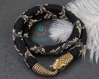 Puede incluir: Un collar con forma de serpiente hecho de cuentas de semillas negras, doradas y moradas iridiscentes. La pieza presenta una cabeza de serpiente y un cierre de cola en tono dorado metálico. El texto STASIA BEADS beaded jewelry es visible.