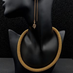 Peut inclure: Un collier tubulaire épais et texturé de couleur dorée avec une finition perlée, accompagné d'une boucle d'oreille unique de type fil avec un petit pendentif en forme de serpent. L'ensemble est présenté sur un buste noir.