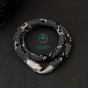 May include: A black and silver beaded snake necklace with a silver snake head clasp. The necklace is coiled in a circle and the snake head is facing the viewer. The necklace is on a black background.