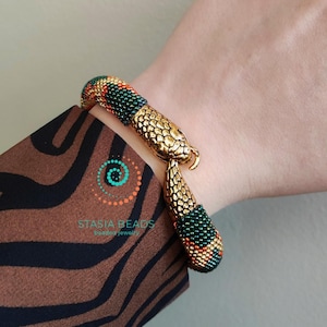 May include: A gold snake bracelet with green, orange, and gold beads. The bracelet is on a person's wrist. The text "STASIA BEADS beaded jewelry" is visible on the image.