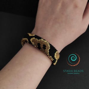 May include: A black and gold beaded bracelet with a leopard print pattern. The bracelet is displayed on a wrist, highlighting the detailed beadwork. The jewellery is from Stasia Beads.