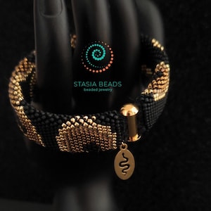 May include: A black and gold beaded bracelet with a leopard print pattern. The bracelet has a gold clasp and a small gold charm with a snake design. The text "STASIA BEADS beaded jewelry" is visible.