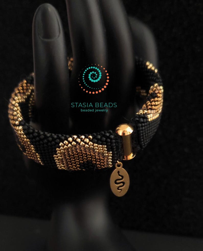May include: A black and gold beaded bracelet with a leopard print pattern. The bracelet has a gold clasp and a small gold charm with a snake design. The text "STASIA BEADS beaded jewelry" is visible.