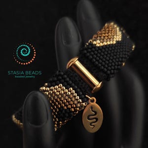 May include: Black beaded bracelet with gold detailing and a gold clasp. The bracelet has a repeating pattern of black and gold beads. A gold charm with a snake design hangs from the bracelet. The text "STASIA BEADS beaded jewelry" is visible.