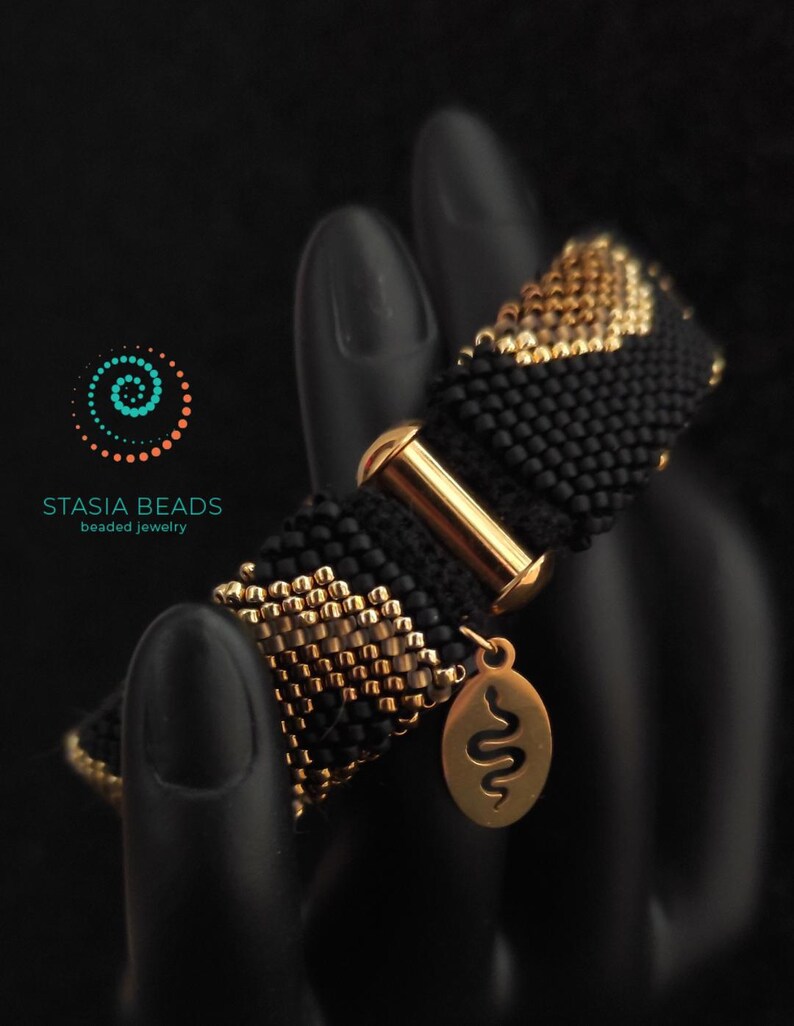May include: Black beaded bracelet with gold detailing and a gold clasp. The bracelet has a repeating pattern of black and gold beads. A gold charm with a snake design hangs from the bracelet. The text "STASIA BEADS beaded jewelry" is visible.