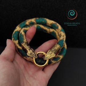 May include: A snake-shaped beaded bracelet featuring a pattern of metallic gold and dark teal beads. The ends of the bracelet are finished with gold-toned metal snake heads that connect to a circular gold ring clasp. Text reads STASIA BEADS.