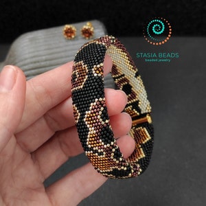 May include: A black, brown, and gold beaded bracelet with a geometric pattern. The bracelet is made with small seed beads and has a gold clasp. The bracelet is shown on a hand.