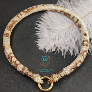 Peut inclure: Un collier de perles blanches et brunes avec des fermoirs en forme de tête de serpent dorés. Le collier est sur un fond noir avec une plume blanche.