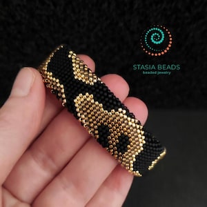 May include: A black and gold beaded bracelet with a leopard print pattern. The bracelet is held in a hand and features the text "STASIA BEADS beaded jewelry" in the upper right corner.