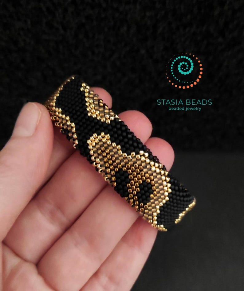 May include: A black and gold beaded bracelet with a leopard print pattern. The bracelet is held in a hand and features the text "STASIA BEADS beaded jewelry" in the upper right corner.