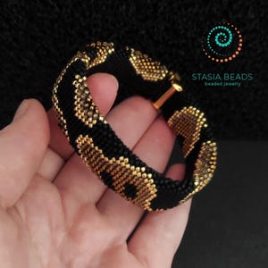 May include: A black and gold beaded bracelet with a repeating pattern. The bracelet is held in a hand against a dark background. The bracelet has a gold clasp and the text "STASIA BEADS beaded jewelry".