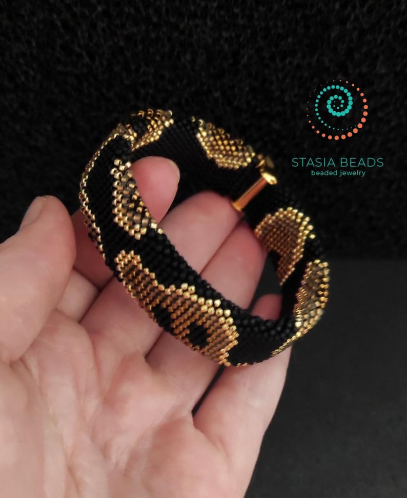 May include: A black and gold beaded bracelet with a repeating pattern. The bracelet is held in a hand against a dark background. The bracelet has a gold clasp and the text "STASIA BEADS beaded jewelry".