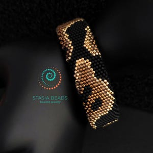 May include: A beaded bracelet with a black and gold pattern. The bracelet is cylindrical and appears to be made of small beads. The design features a repeating pattern of black and gold shapes. The bracelet is displayed against a dark background.