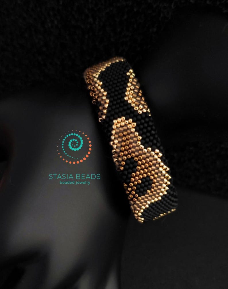 May include: A beaded bracelet with a black and gold pattern. The bracelet is cylindrical and appears to be made of small beads. The design features a repeating pattern of black and gold shapes. The bracelet is displayed against a dark background.