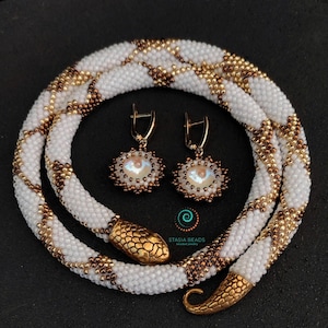 Könnte beinhalten: Weiße und goldene Perlen-Schlangenhalskette mit passenden Ohrringen. Die Halskette hat einen goldenen Schlangenkopfverschluss und die Ohrringe haben irisierende Glasperlen.