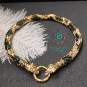 Puede incluir: Un collar de cuentas doradas con un diseño de serpiente y un cierre dorado. Las cuentas son verdes y doradas y tienen un acabado brillante. El collar está sobre un fondo de pluma blanca.