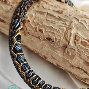 May include: A beaded necklace with a black, gold, and blue geometric pattern. The necklace is coiled on a piece of wood.