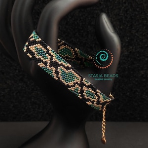 May include: A beaded bracelet with a snake scale pattern in black, teal, and gold. The bracelet is displayed on a black hand-shaped jewelry holder. The bracelet has a gold chain and clasp. The brand name "STASIA BEADS" is visible.