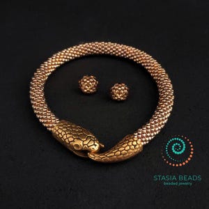 May include: A gold-toned beaded snake-shaped necklace and matching stud earrings set. The snake's head and tail are detailed with textured scales. The jewelry is displayed on a black surface. The brand logo "STASIA BEADS beaded jewelry" is in the bottom right corner.