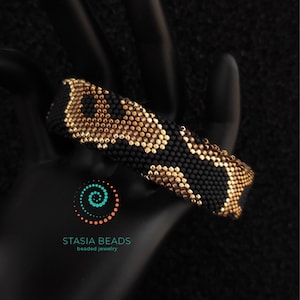 May include: A black and gold beaded bracelet with a leopard print pattern. The bracelet is made of small beads and is shown on a black surface. The Stasia Beads logo is visible.
