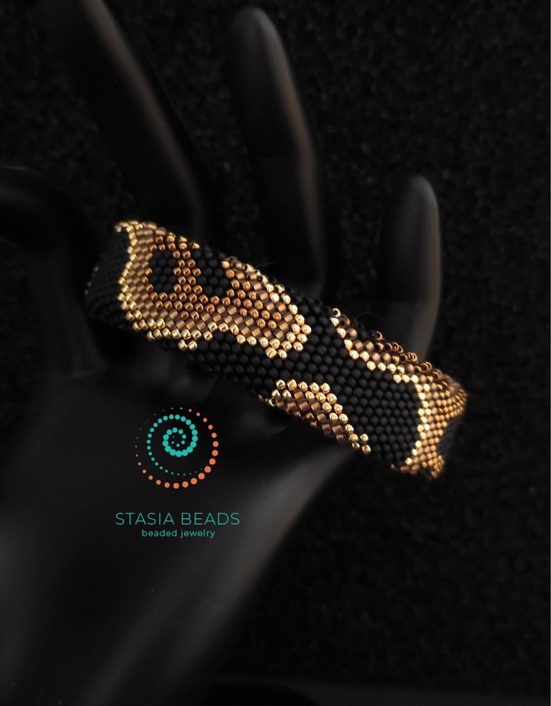 May include: A black and gold beaded bracelet with a leopard print pattern. The bracelet is made of small beads and is shown on a black surface. The Stasia Beads logo is visible.