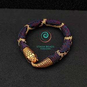 May include: A beaded snake bracelet with a gold snake head clasp. The bracelet is made with black, purple, and gold beads. The bracelet is a statement piece and would be a great addition to any jewelry collection.