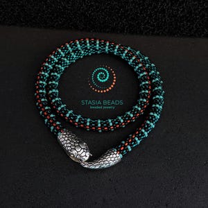 May include: A beaded necklace with a snake design. The necklace features a pattern of black, teal, and red beads. The clasp is a silver-colored snake head. The necklace is displayed on a black surface. The text "STASIA BEADS beaded jewelry" is also visible.