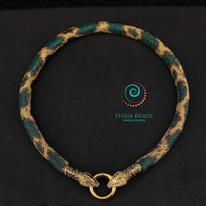 May include: A gold and green beaded necklace with a snake design and gold snake head clasps. The necklace is on a black background.  Stasia Beads beaded jewelry.