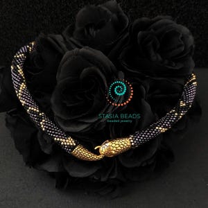 Peut inclure: Un collier de serpent en perles noires et dorées avec un fermoir en forme de serpent doré. Le collier est sur un fond noir avec des roses noires.