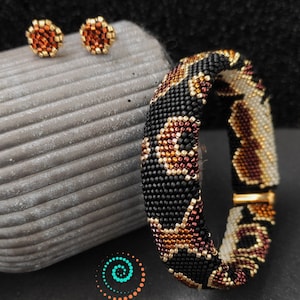 Op de afbeelding: Een paar kleine, ronde oorbellen met een zwart, bruin en gouden parelmotief. Een grotere parel armband met een vergelijkbaar ontwerp is ook afgebeeld. De armband is gemaakt van zwarte, bruine en gouden kralen en heeft een geometrisch patroon.