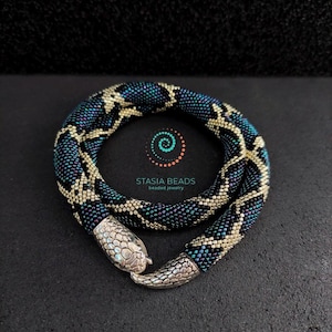 May include: A beaded necklace in a snake design. The necklace features a pattern of teal, black, and gold beads. The clasp is silver-toned and shaped like a snake's head. The necklace is displayed on a dark surface. The text "STASIA BEADS beaded jewelry" is visible.