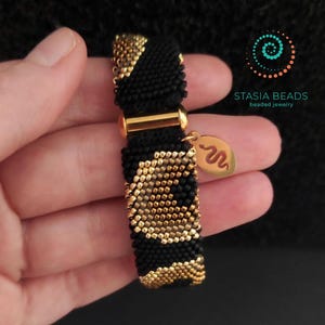 May include: A black and gold beaded bracelet with a gold clasp and a gold charm. The bracelet has a repeating pattern of black and gold beads. The charm features a snake design. The brand name "STASIA BEADS beaded jewelry" is visible.