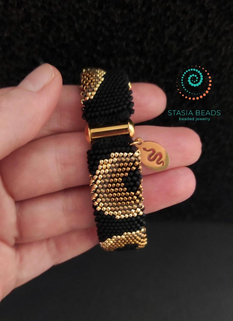 May include: A black and gold beaded bracelet with a gold clasp and a gold charm. The bracelet has a repeating pattern of black and gold beads. The charm features a snake design. The brand name "STASIA BEADS beaded jewelry" is visible.