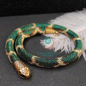 May include: A green and gold beaded snake necklace with a gold snake head charm. The necklace is made of small, iridescent beads and has a textured, scaly appearance. The necklace is coiled and resting on a white feather.