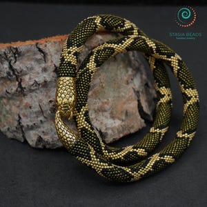 Puede incluir: Un collar o pulsera largo y flexible de cuentas con diseño de serpiente, con un cierre de metal dorado en forma de cabeza y cola. El cuerpo está hecho de pequeñas cuentas negras y doradas en un patrón de rombos. El texto dice STASIA BEADS.
