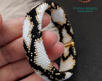 Op de afbeelding: Een buisvormige kralenarmband met een geometrisch patroon van witte, zwarte en metallic gouden kraaltjes. Het ontwerp bestaat uit herhalende chevronvormen. De tekst STASIA BEADS beaded jewelry is zichtbaar in de rechterbovenhoek.