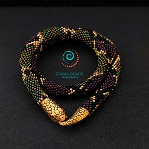 May include: A coiled snake-shaped necklace crafted from small black, green, and purple beads, featuring a polished gold-tone metal head and tail clasp. The centre of the coil displays the text STASIA BEADS beaded jewellery with a spiral logo.
