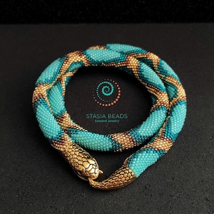 May include: A turquoise and gold beaded snake necklace. The necklace is made of small, round beads in a pattern of turquoise and gold. The snake head clasp is gold-coloured and detailed. The necklace is a unique piece of beaded jewellery.