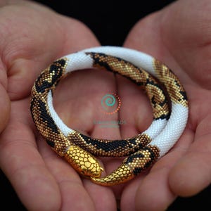 May include: A coiled snake necklace crafted from tiny beads in a pattern of white, black, and metallic bronze. The snake features a textured gold-toned metal head and tail tip. A small logo with a spiral design and the text STARBEADS is visible.
