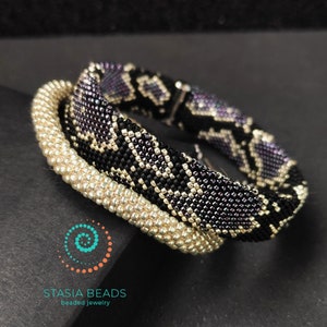 May include: Three beaded bracelets stacked on top of each other. The top bracelet is black with a silver and purple snake skin pattern. The middle bracelet is a solid silver color. The bottom bracelet is a solid gold color. The bracelets are all made of small beads.
