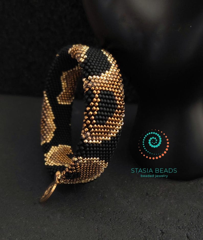 May include: A beaded bracelet against a black backdrop. The bracelet features a black base with a gold leopard print design. It has a gold charm and is labelled "STASIA BEADS beaded jewelry".