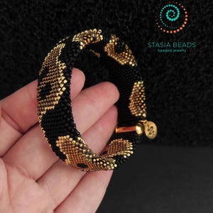 May include: A black and gold beaded bracelet with a leopard print pattern. The bracelet is held in a hand, highlighting the detailed beadwork and the gold-coloured clasp. The brand name "STASIA BEADS" is displayed.