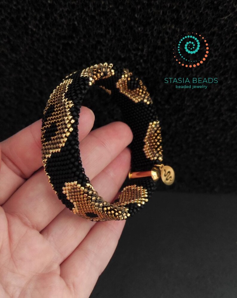 May include: A black and gold beaded bracelet with a leopard print pattern. The bracelet is held in a hand, highlighting the detailed beadwork and the gold-coloured clasp. The brand name "STASIA BEADS" is displayed.