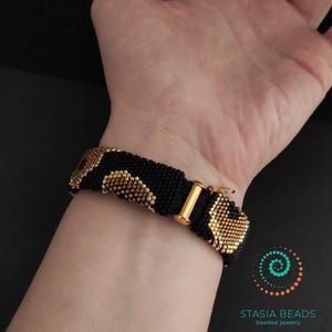 May include: Black beaded bracelet with gold-coloured accents and a gold-tone clasp. The bracelet features a repeating pattern of gold beads on a black background. The bracelet is shown on a wrist.