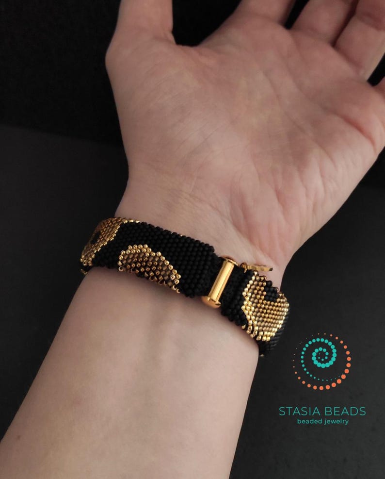 May include: Black beaded bracelet with gold-coloured accents and a gold-tone clasp. The bracelet features a repeating pattern of gold beads on a black background. The bracelet is shown on a wrist.
