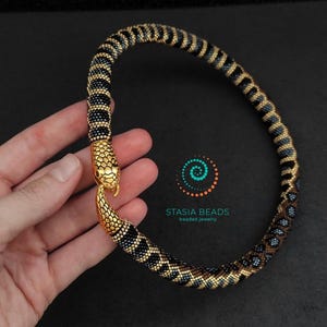 May include: A beaded necklace featuring a flexible, tubular body with a repeating pattern of black, gold, and blue beads. The necklace ends in a gold-toned metal snake head clasp. The text STASIA BEADS beaded jewelry is visible in the center.