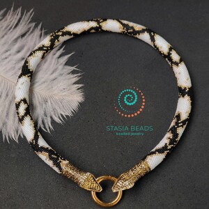 May include: A white and black beaded necklace with a gold snake design and a gold clasp. The necklace is on a black background with a white feather.