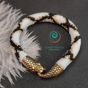 May include: A white, black and gold beaded snake bracelet with gold snake head clasps. The bracelet is on a grey surface with a white feather in the background. The text "Stasia Beads beaded jewellery" is visible in the image.