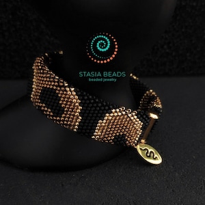 May include: Black and gold beaded bracelet with a snake design. The bracelet is made of small, round beads and features a gold-colored clasp and charm. The charm has a snake symbol and the text "STASIA BEADS beaded jewelry".